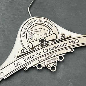 May include: Silver graduation hanger with a black engraved design. The design includes a graduation cap, diploma, and the text "University of Adelaide, Dr. Pamela Crossman PhD, Graduation 12/19/2023".