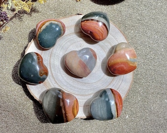 May include: A collection of polished heart-shaped stones in shades of green, brown, and red arranged on a wooden slice. A large, patterned seashell sits above the stones. Dried flowers are in the background. The stones are likely used for decorative purposes.