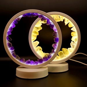 May include: Two white, circular lamps with a ring of purple and yellow crystals. The lamps emit a soft glow, and the crystals are illuminated. The lamps sit on white bases, against a black background.