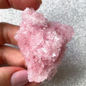 May include: A cluster of translucent, light pink crystalline mineral formations with a jagged, geometric surface texture. The specimen features numerous small, sharp-edged cubic crystals that catch the light, creating a sparkling appearance across the entire structure.