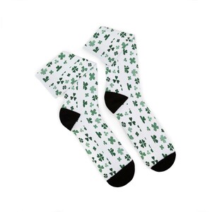 Puede incluir: Un par de calcetines blancos con un estampado de tréboles verdes. Los calcetines tienen talones y punteras negras. Estos calcetines son probablemente para el Día de San Patricio.