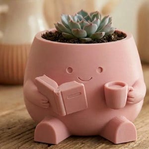 May include: A pink, whimsical planter shaped like a character, holding a book and a mug. The planter has a smiling face and is filled with soil and a succulent plant. The overall design is charming and decorative.