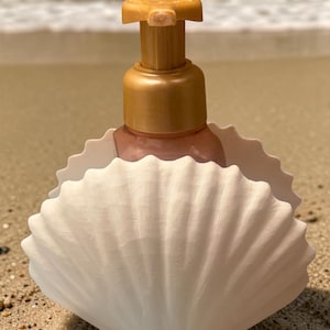 May include: A decorative soap dispenser with a gold-colored pump and a white, seashell-shaped holder. The dispenser is filled with a pink liquid and sits on a sandy beach with the ocean in the background.
