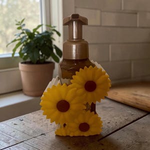May include: A brown soap dispenser decorated with three yellow sunflowers with brown centers. The dispenser is on a wooden surface near a window and a potted plant.
