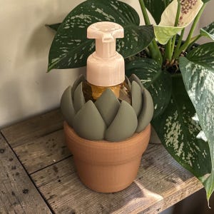 May include: A decorative soap dispenser featuring a light pink pump and a clear bottle. The bottle is nestled in a sage green succulent-shaped holder, which sits inside a terracotta-colored pot. The dispenser is placed on a wooden surface.