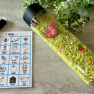 May include: A clear plastic sensory bottle filled with yellow and green rice, small farm-themed charms, and a black lid. A laminated "I Spy" game card and a dry-erase marker are next to the bottle. The card features farm-related images.