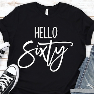 May include: Black t-shirt with white text that says "Hello Sixty". The shirt is laid out on a white wood background with a pair of black trainers and blue jeans.