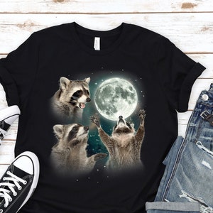 May include: Black t-shirt featuring a graphic of three raccoons looking up at a full moon. The raccoons are in a playful pose, with one reaching up towards the moon.