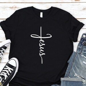 May include: Black t-shirt with a white cross and the word "Jesus" written vertically on it. The shirt is laid out on a white wooden surface with a pair of black sneakers and blue jeans.