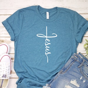 K&ouml;nnte beinhalten: Ein t&uuml;rkisfarbenes T-Shirt mit einem wei&szlig;en Kreuz und dem Wort "Jesus", das vertikal darauf geschrieben ist. Das Shirt liegt auf einer Holzoberfl&auml;che mit einem Paar wei&szlig;en Turnschuhen und einer Jeans-Shorts.