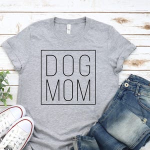 May include: A grey t-shirt with the words "DOG MOM" in a black square, alongside blue denim jeans and white canvas trainers. The casual outfit is displayed on a light wood-effect surface, suggesting a relaxed, everyday style.