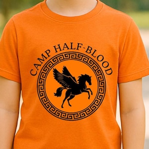May include: Orange t-shirt with a black graphic of a winged horse inside a circular design, with the words "CAMP HALF-BLOOD" in a semi-circle above the graphic. The shirt is worn by a young person with blonde hair and blue eyes.