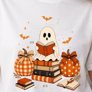 May include: A white short-sleeved t-shirt featuring a graphic print of a ghost reading a book while sitting on a stack of books. The design includes two pumpkins, one with a plaid pattern and one with polka dots, surrounded by small bats and spiders.