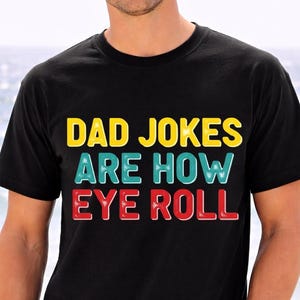 Peut inclure: T-shirt noir avec l'inscription "DAD JOKES ARE HOW EYE ROLL" en jaune, bleu sarcelle et rouge. La personne porte un jean bleu et se tient sur une plage.