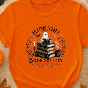 May include: An orange short-sleeved t-shirt featuring a graphic of a ghost reading a book atop a stack of books. The text reads MIDNIGHT BOOK SOCIETY, READ MORE, STAY CURIOUS, LIVE IN STORIES. The design includes a candle, a mug, and bats.