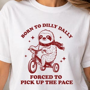 May include: A white short-sleeved t-shirt featuring a dark red graphic of a sloth wearing a scarf and riding a tricycle. The text above the sloth reads BORN TO DILLY DALLY and the text below the sloth reads FORCED TO PICK UP THE PACE.