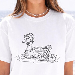 May include: White t-shirt featuring a black and white illustration of a goose wearing a bonnet and scarf, with two ducklings in the water. The goose is swimming in a pond. The shirt is a casual style.