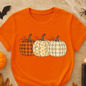 May include: A bright orange short-sleeved t-shirt featuring a graphic print of three pumpkins in a row. The first pumpkin has a dark brown plaid pattern, the middle one has a floral print, and the third has a light beige and white checkered pattern.
