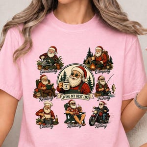 May include: A light pink t-shirt featuring a central graphic of Santa Claus holding a coffee mug with the text "LIVING MY BEST LIFE". Surrounding the centre are smaller illustrations of Santa gardening, camping, reading, fishing, cooking, relaxing, and riding a motorcycle, each labelled with its respective activity.