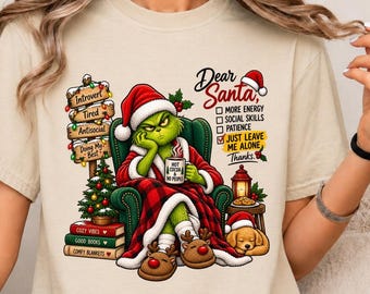Könnte beinhalten: Ein beiges T-Shirt mit einer Grafik einer grünen Figur mit Weihnachtsmütze, die in einem grünen Sessel mit einer rot karierten Decke sitzt. Die Grafik zeigt zudem ein Wegweiser-Schild, Bücher, einen Weihnachtsbaum, einen Hund und den Text Dear Santa.
