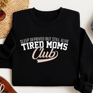 May include: Black sweatshirt with the text "SLEEP DEPRIVED BUT STILL ALIVE TIRED MOMS Club" in white and pink lettering. The sweatshirt is a crew neck style.