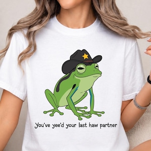 May include: A white short-sleeved t-shirt featuring a graphic of a green frog wearing a black cowboy hat with a yellow star badge. Below the frog, the text reads, "You've yee'd your last haw partner" in a simple black sans-serif font.