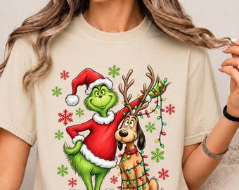 Könnte beinhalten: Ein beiges Kurzarm-T-Shirt mit einem Grafikdruck einer grünen Figur im roten Weihnachtsmannkostüm neben einem braunen Hund mit Geweih. Der Hund ist mit bunten Weihnachtslichtern geschmückt und das Design enthält rote und grüne Schneeflocken.