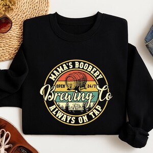 May include: Black sweatshirt with a circular graphic that reads "Mama's Boobery Brewing Co" in a vintage style. The design features a beer barrel, sunset colors, and the words "Always on Tap".