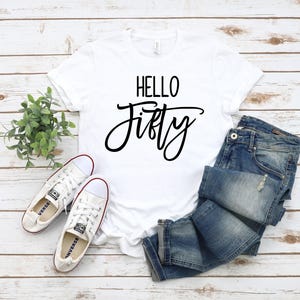 May include: White t-shirt with black text that says "Hello Fifty". The shirt is laying on a white wooden surface with a pair of white sneakers, a potted plant, and a pair of blue jeans.