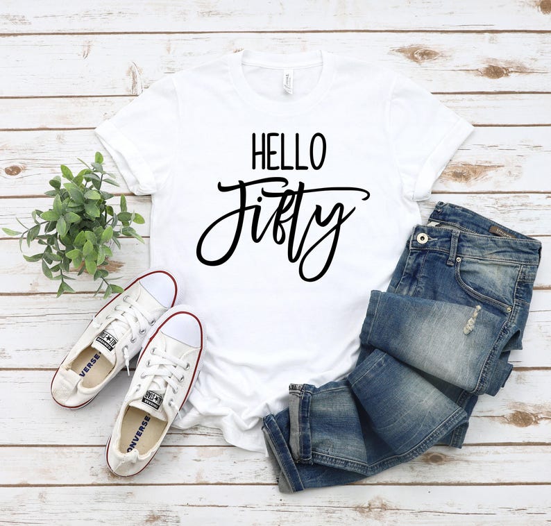 May include: White t-shirt with black text that says "Hello Fifty". The shirt is laying on a white wooden surface with a pair of white sneakers, a potted plant, and a pair of blue jeans.