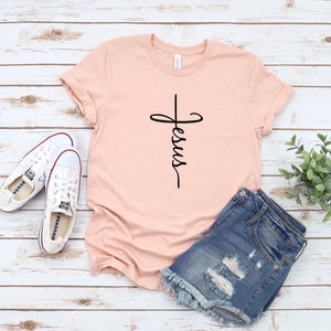 K&ouml;nnte beinhalten: Ein hellrosa T-Shirt mit einem schwarzen Kreuz und dem Wort "Jesus", das vertikal darauf geschrieben ist. Das Shirt liegt auf einer wei&szlig;en Holzoberfl&auml;che mit einem Paar wei&szlig;en Turnschuhen und einer blauen Jeans-Shorts.