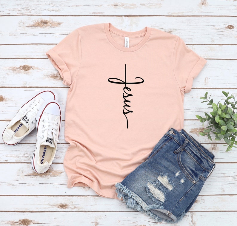 K&ouml;nnte beinhalten: Ein hellrosa T-Shirt mit einem schwarzen Kreuz und dem Wort "Jesus", das vertikal darauf geschrieben ist. Das Shirt liegt auf einer wei&szlig;en Holzoberfl&auml;che mit einem Paar wei&szlig;en Turnschuhen und einer blauen Jeans-Shorts.