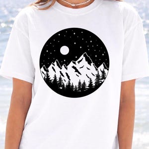 A woman wearing a white t-shirt with a black and white graphic design depicting a mountain landscape with a full moon and stars in the night sky.