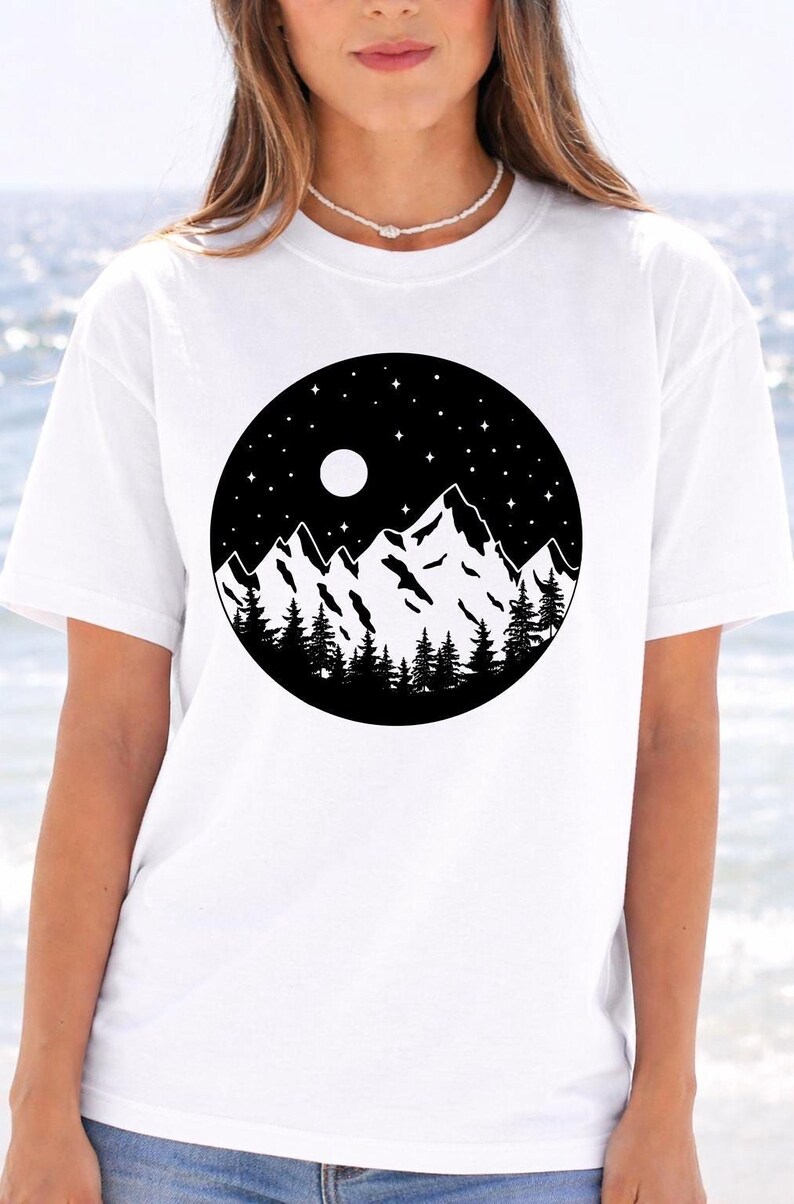 A woman wearing a white t-shirt with a black and white graphic design depicting a mountain landscape with a full moon and stars in the night sky.
