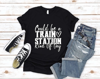 May include: Black t-shirt with the text "Could be a Train Station Kind Of Day" in white. The shirt is paired with blue jeans, black sneakers, and a watch.