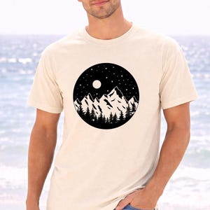 A man wearing a white t-shirt with a graphic design of mountains and a night sky.