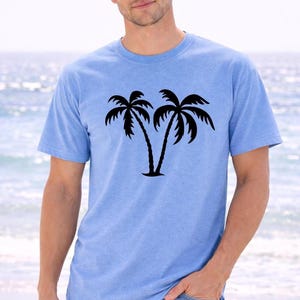 A man wearing a light blue t-shirt with a graphic of two palm trees stands on a beach, with the ocean visible in the background.