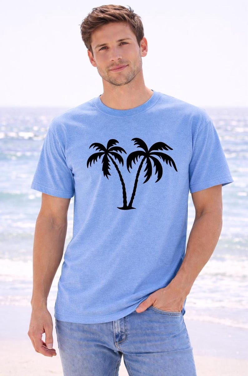 A man wearing a light blue t-shirt with a graphic of two palm trees stands on a beach, with the ocean visible in the background.