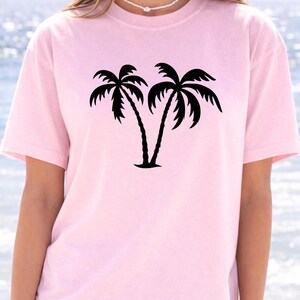 A woman wearing a pink t-shirt with a graphic of two palm trees printed on it, standing in front of a body of water.