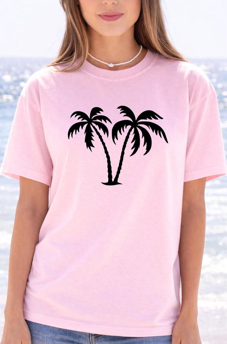A woman wearing a pink t-shirt with a graphic of two palm trees printed on it, standing in front of a body of water.