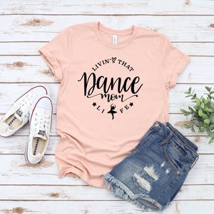 A pink t-shirt with a black text design, a pair of denim shorts, and a pair of white sneakers are laid out on a wooden surface.