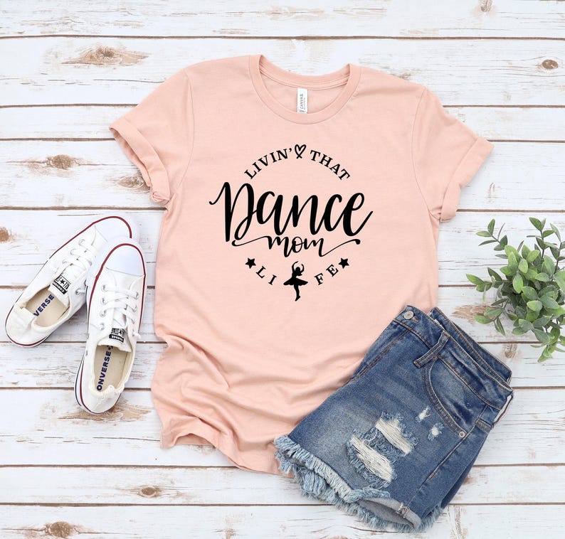 A pink t-shirt with a black text design, a pair of denim shorts, and a pair of white sneakers are laid out on a wooden surface.