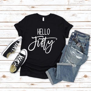 May include: Black t-shirt with white text that says "HELLO FIFTY" on a white wooden background. The t-shirt is laying on top of a pair of blue jeans and two black sneakers.