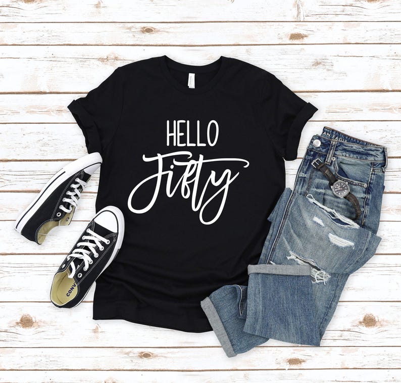 May include: Black t-shirt with white text that says "HELLO FIFTY" on a white wooden background. The t-shirt is laying on top of a pair of blue jeans and two black sneakers.
