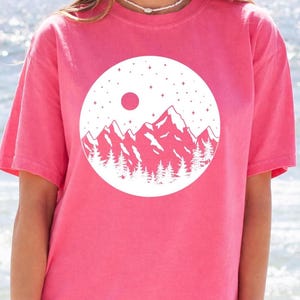 A woman wearing a pink t-shirt with a white graphic design of mountains and a red moon on it, standing in front of a body of water.