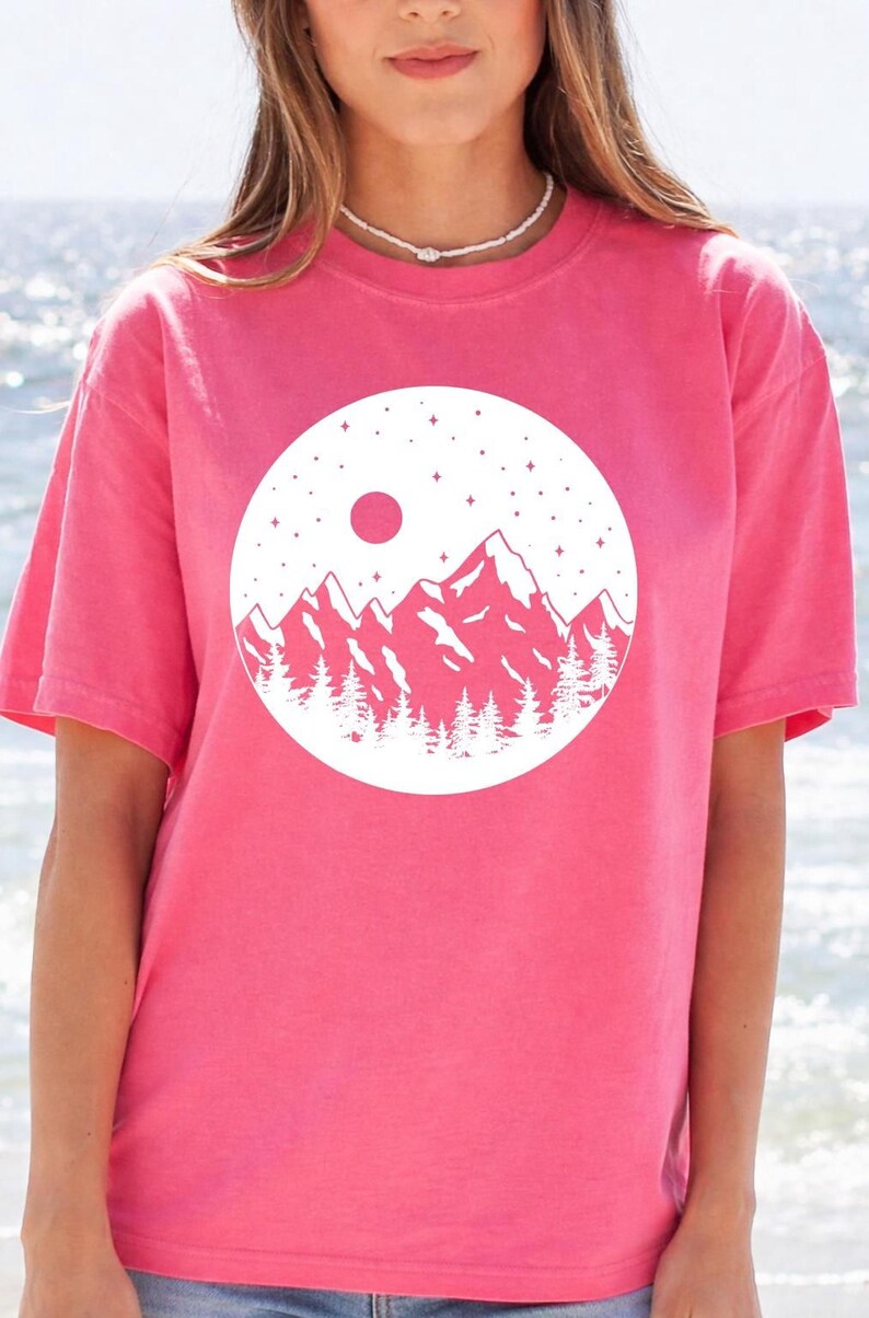 A woman wearing a pink t-shirt with a white graphic design of mountains and a red moon on it, standing in front of a body of water.