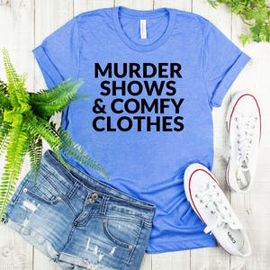a casual outfit consisting of a blue t-shirt with the text "MURDER SHOWS & COMfy CLOTHES", denim shorts, and white sneakers, all laid out on a wooden surface.