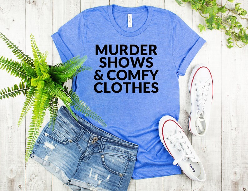 a casual outfit consisting of a blue t-shirt with the text "MURDER SHOWS & COMfy CLOTHES", denim shorts, and white sneakers, all laid out on a wooden surface.