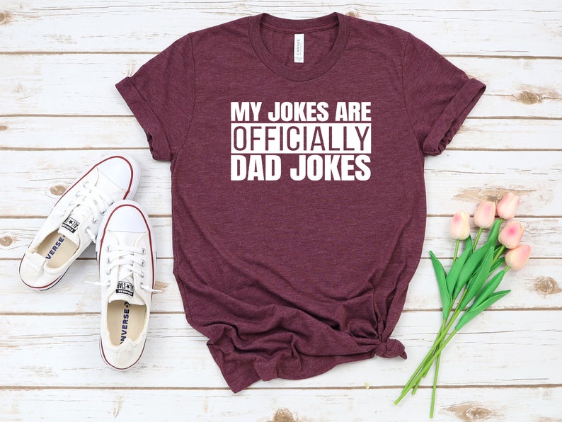 K&ouml;nnte beinhalten: Ein maroonfarbenes T-Shirt mit dem Text "My Jokes Are Officially Dad Jokes" in Wei&szlig;.