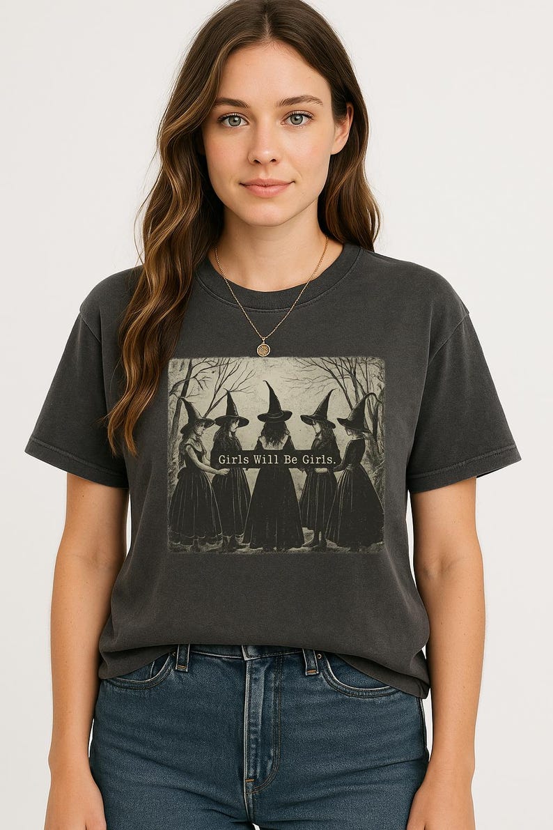 May include: Dark gray t-shirt featuring a graphic of five witches in black dresses and hats, with the text "Girls Will Be Girls." The model is wearing a gold necklace and blue jeans.